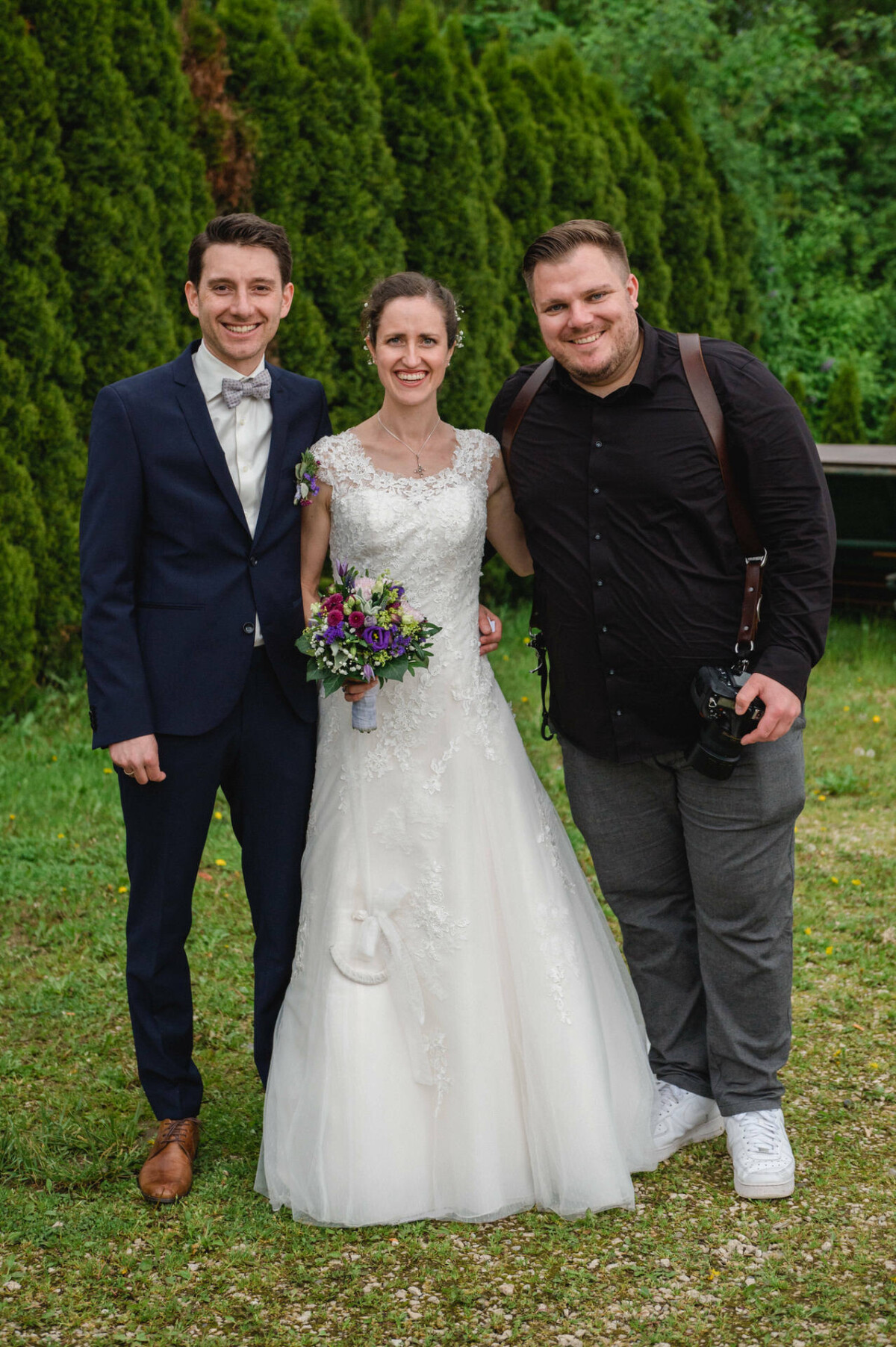 Braut und Bräutigam stehen neben einem Fotografen im Freien, im Hintergrund ist Grün. Die Braut hält einen Blumenstrauß und alle lächeln.