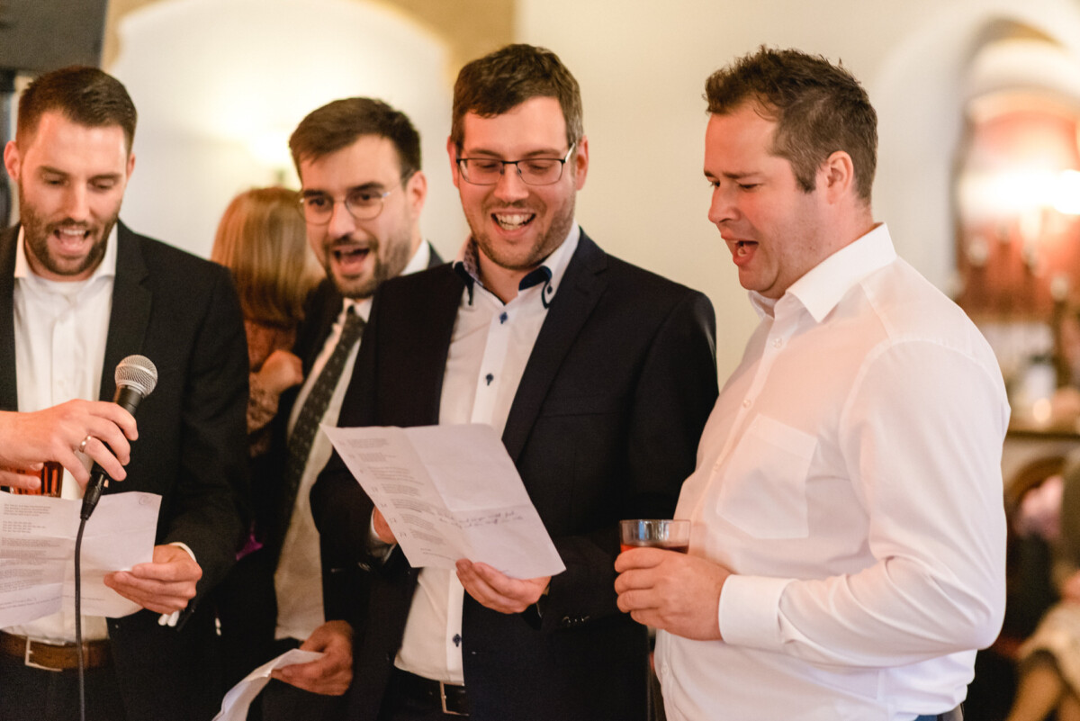 Vier Männer in Abendgarderobe singen zusammen und halten Papiere und ein Mikrofon in den Händen. Ein Mann hält ein Getränk in der Hand.