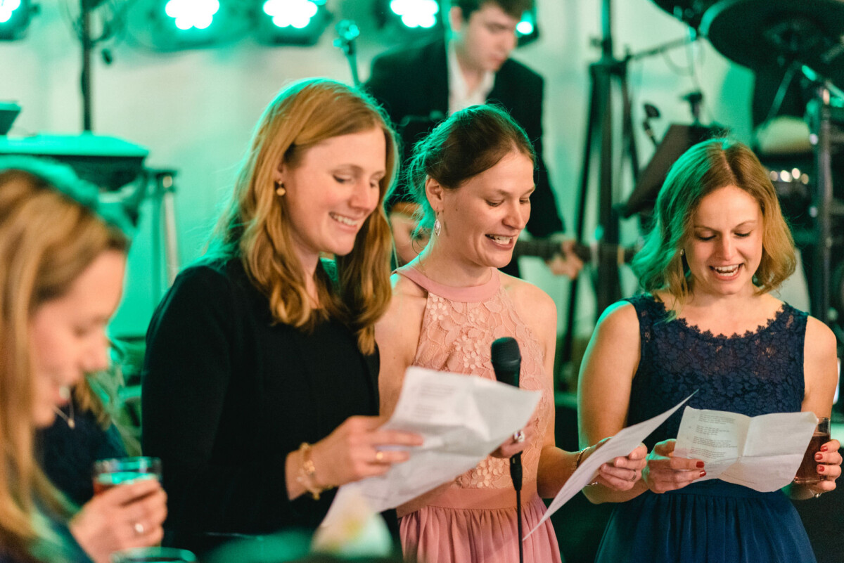 Drei Frauen stehen zusammen, lächeln und halten Papiere, während sie bei einer Indoor-Veranstaltung in ein Mikrofon sprechen oder singen.