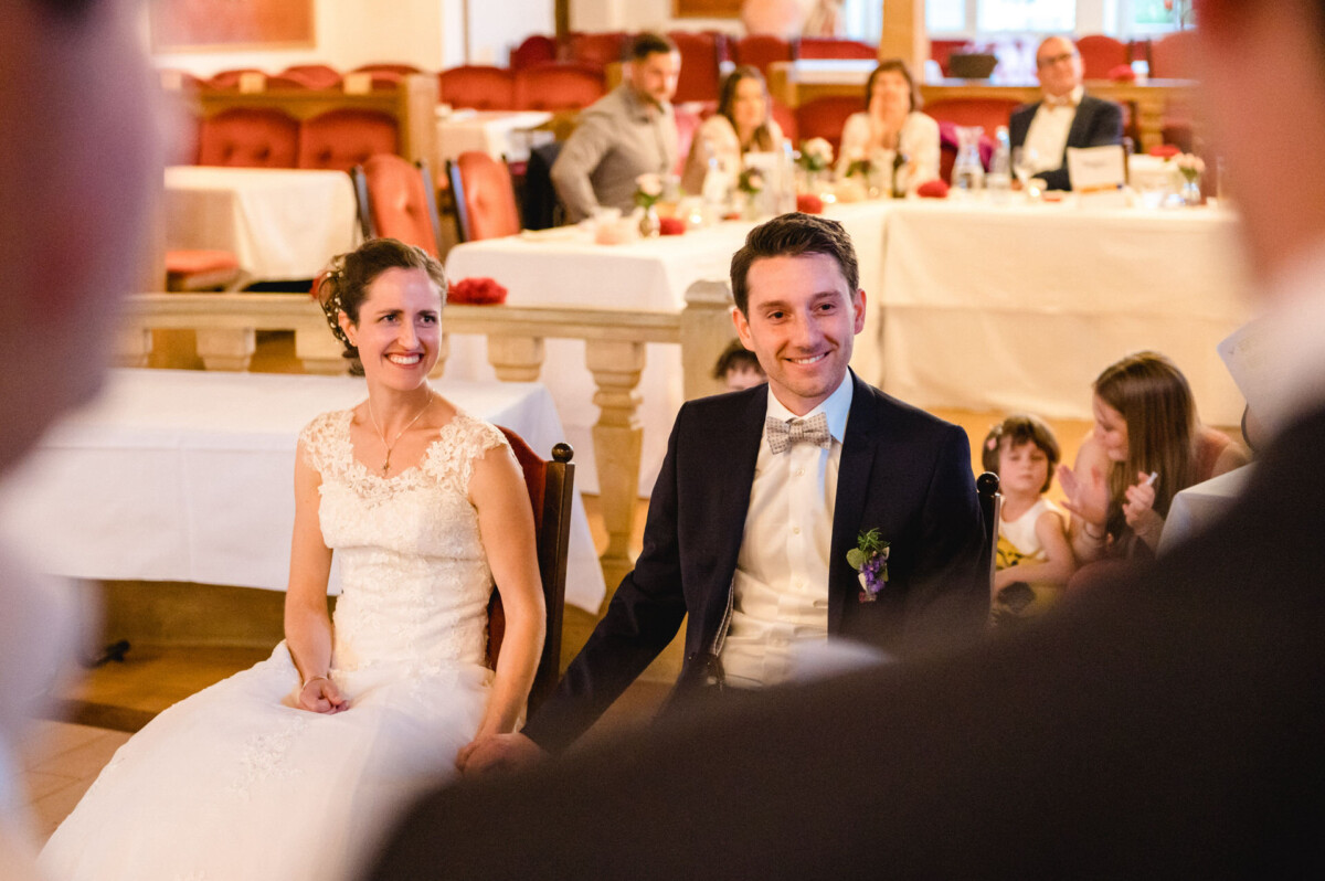 Eine Braut und ein Bräutigam sitzen lächelnd bei einem Hochzeitsempfang zusammen, im Hintergrund sitzen die Gäste.