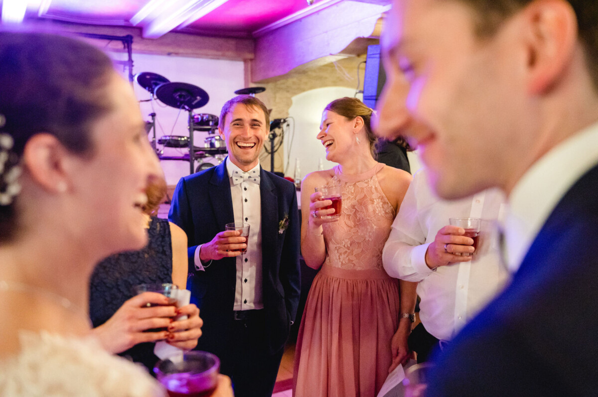 Eine Gruppe von Menschen in Abendgarderobe unterhält sich und lacht, hält Getränke in der Hand und im Hintergrund sind Musikinstrumente zu sehen.