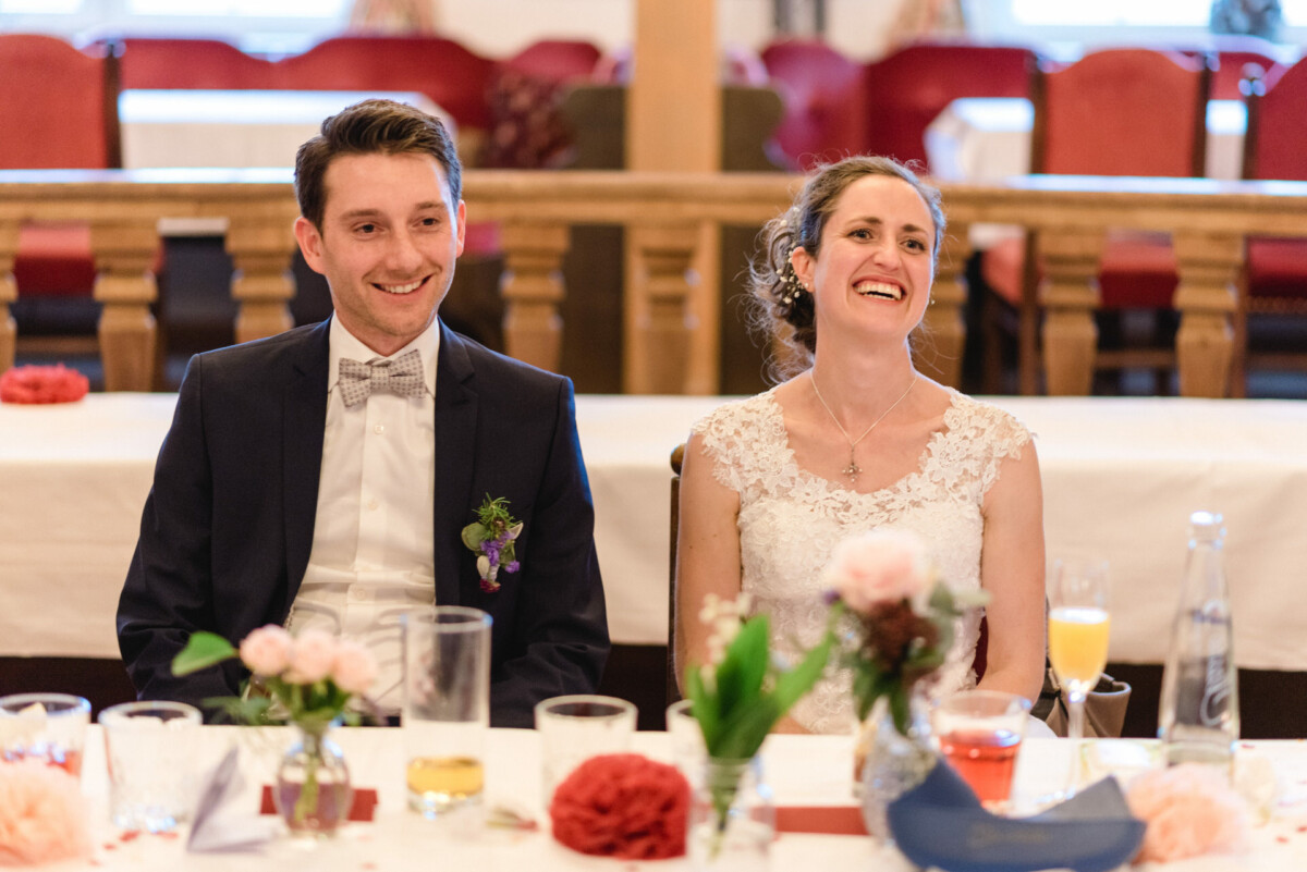 Ein Paar im Hochzeitskleid sitzt lächelnd an einem gedeckten Tisch.
