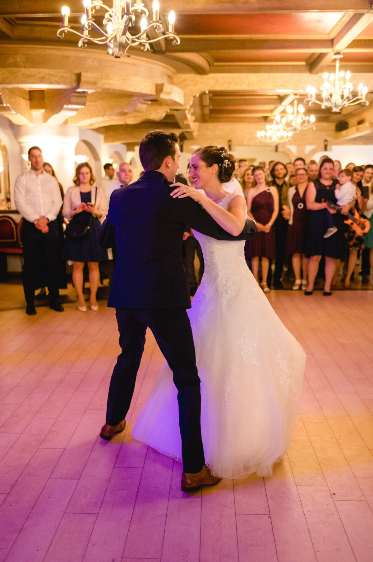 Ein Paar tanzt zusammen in einem Ballsaal. Die Frau trägt ein weißes Kleid und der Mann einen dunklen Anzug. Im Hintergrund schaut eine Gruppe von Menschen zu und über ihnen hängen Kronleuchter.