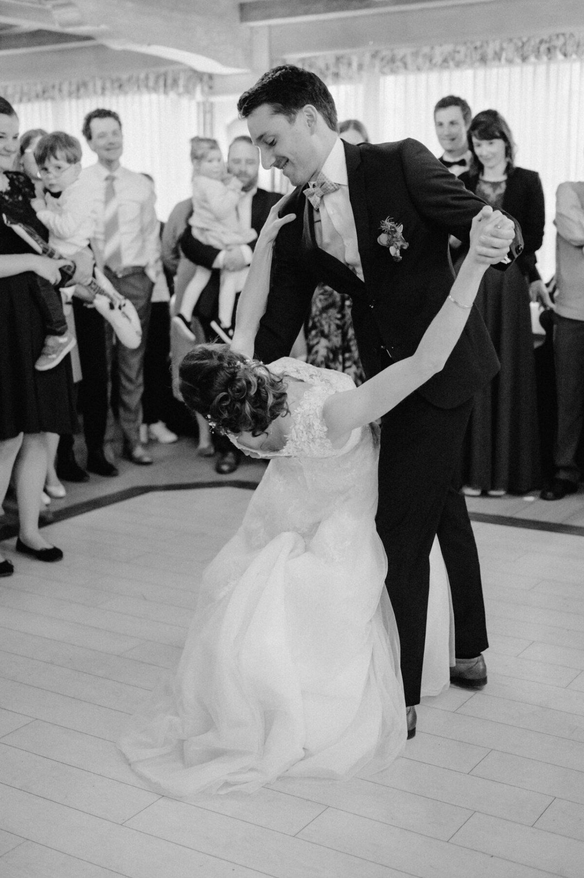 Ein Paar tanzt in einem Ballsaal, umgeben von Gästen, während der Bräutigam die Braut ins Wasser taucht. Die Szene ist in Schwarzweiß festgehalten.