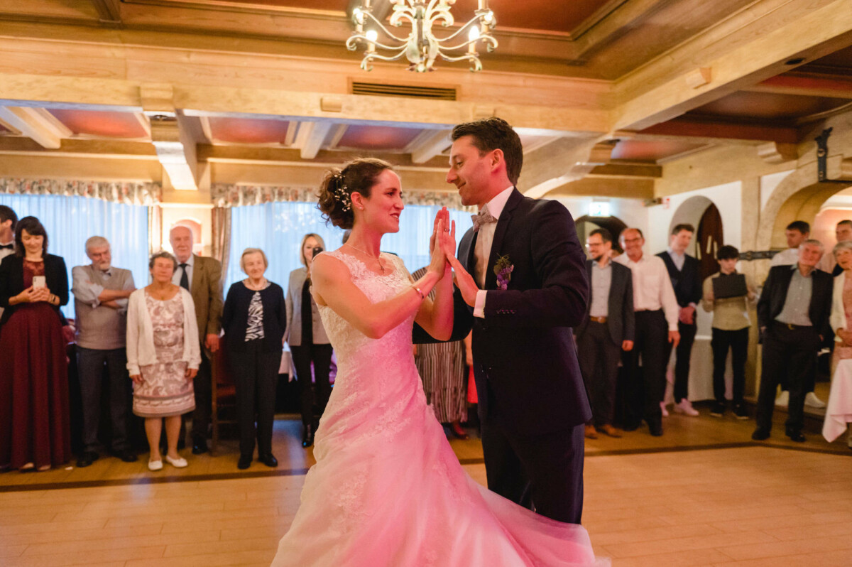 Braut und Bräutigam tanzen in einem warm beleuchteten Raum mit einem Kronleuchter darüber, umgeben von stehenden Gästen, die ihnen zuschauen.