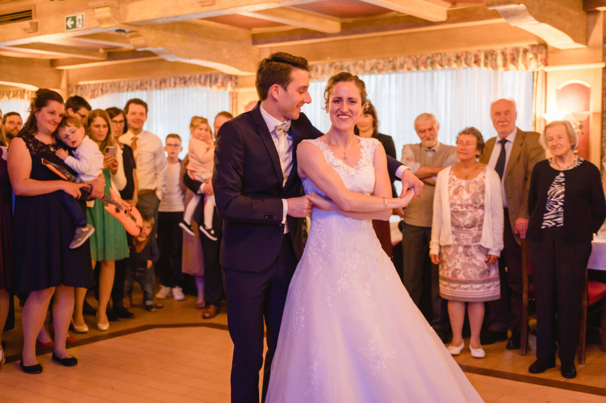 Ein Brautpaar tanzt umgeben von Gästen und mit einem kleinen Jungen in den Armen eines Zuschauers in einem warm beleuchteten Raum.