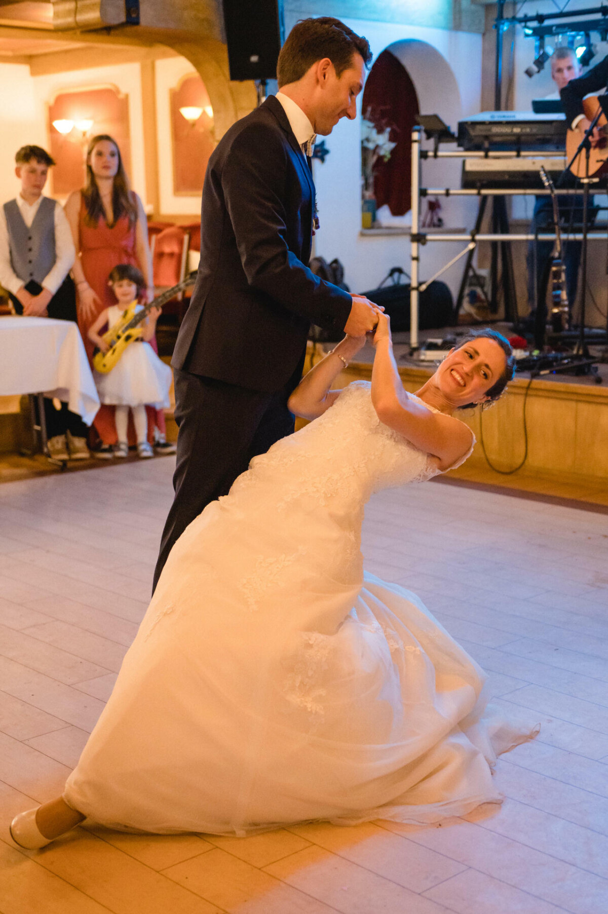 Ein Paar in Abendgarderobe tanzt, wobei der Mann die Frau hält, während sie sich zurücklehnt. Ein Junge und ein Mädchen sitzen im Hintergrund an einem Tisch und auf einer Bühne spielt eine Band.