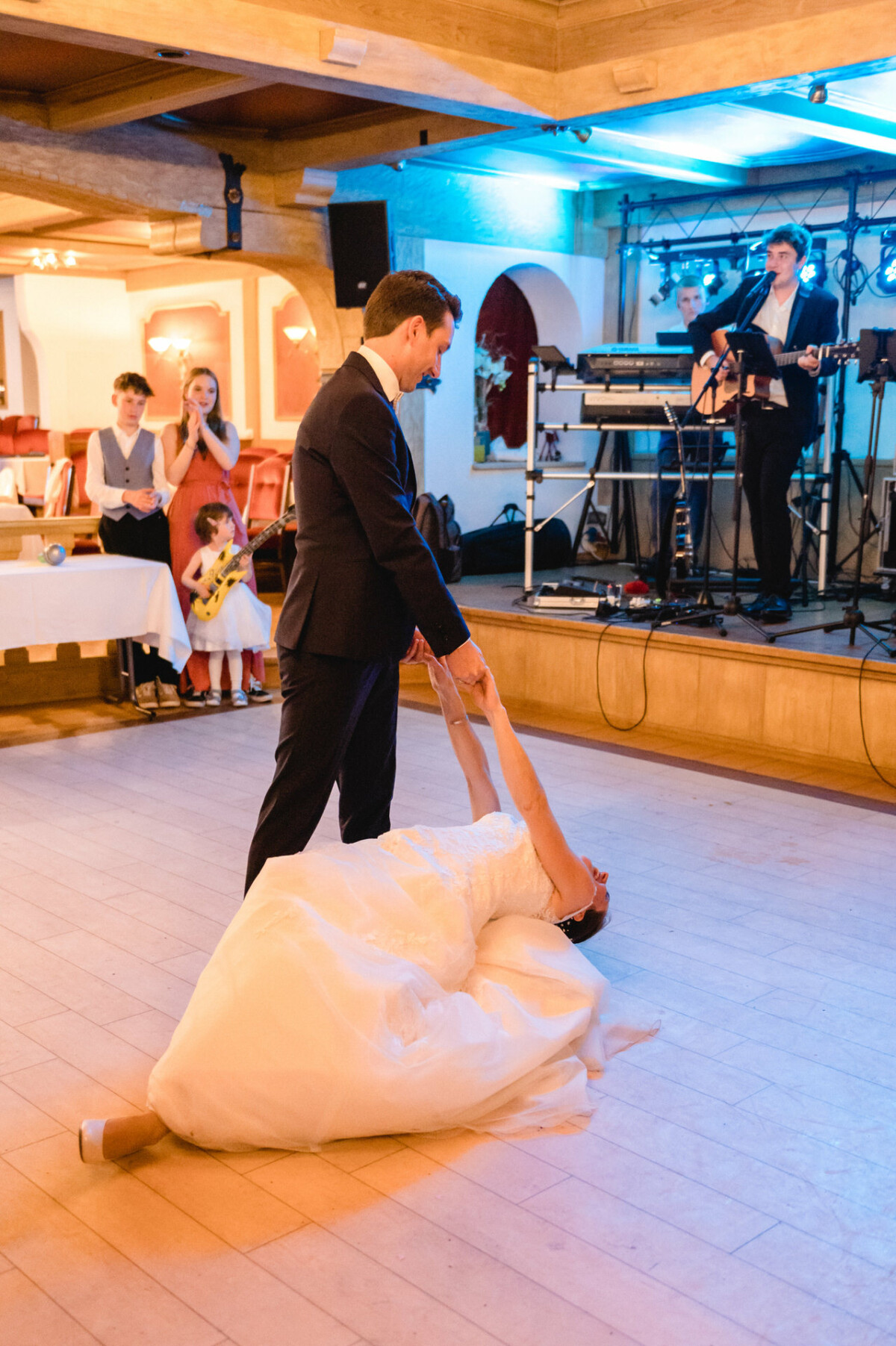 Braut und Bräutigam tanzen in einem Bankettsaal, im Hintergrund spielt eine Live-Band und die Gäste sitzen in der Nähe.