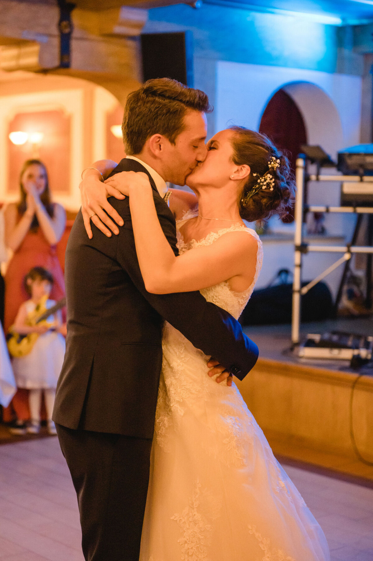 Braut und Bräutigam küssen sich während ihrer Hochzeitsfeier auf der Tanzfläche, während die Gäste zuschauen und im Hintergrund bunte Lichter spielen.