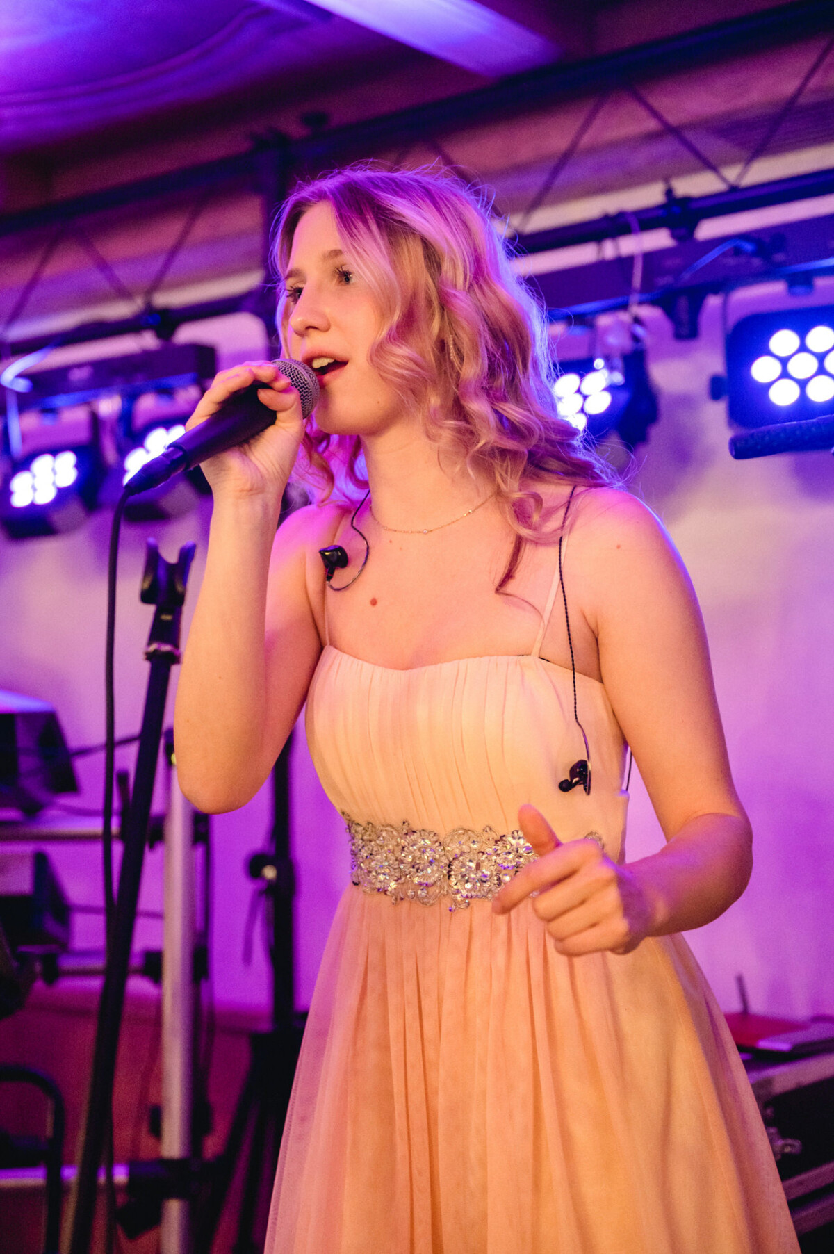 Eine Person in einem rosa Kleid singt auf der Bühne in ein Mikrofon, im Hintergrund sind bunte Lichter zu sehen.