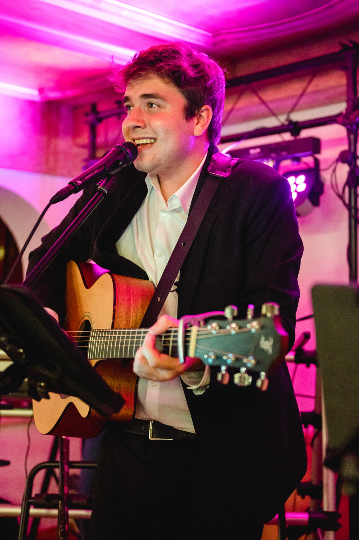 Ein Mann im Anzug singt in ein Mikrofon, während er unter violettem Bühnenlicht eine Akustikgitarre spielt.