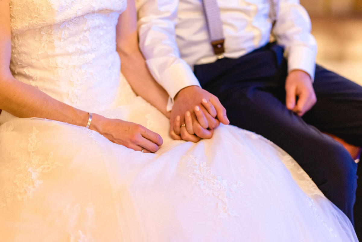 Ein Paar in Abendgarderobe sitzt Händchen haltend da; die Frau trägt ein weißes Spitzenkleid und der Mann ein weißes Hemd mit Hosenträgern.