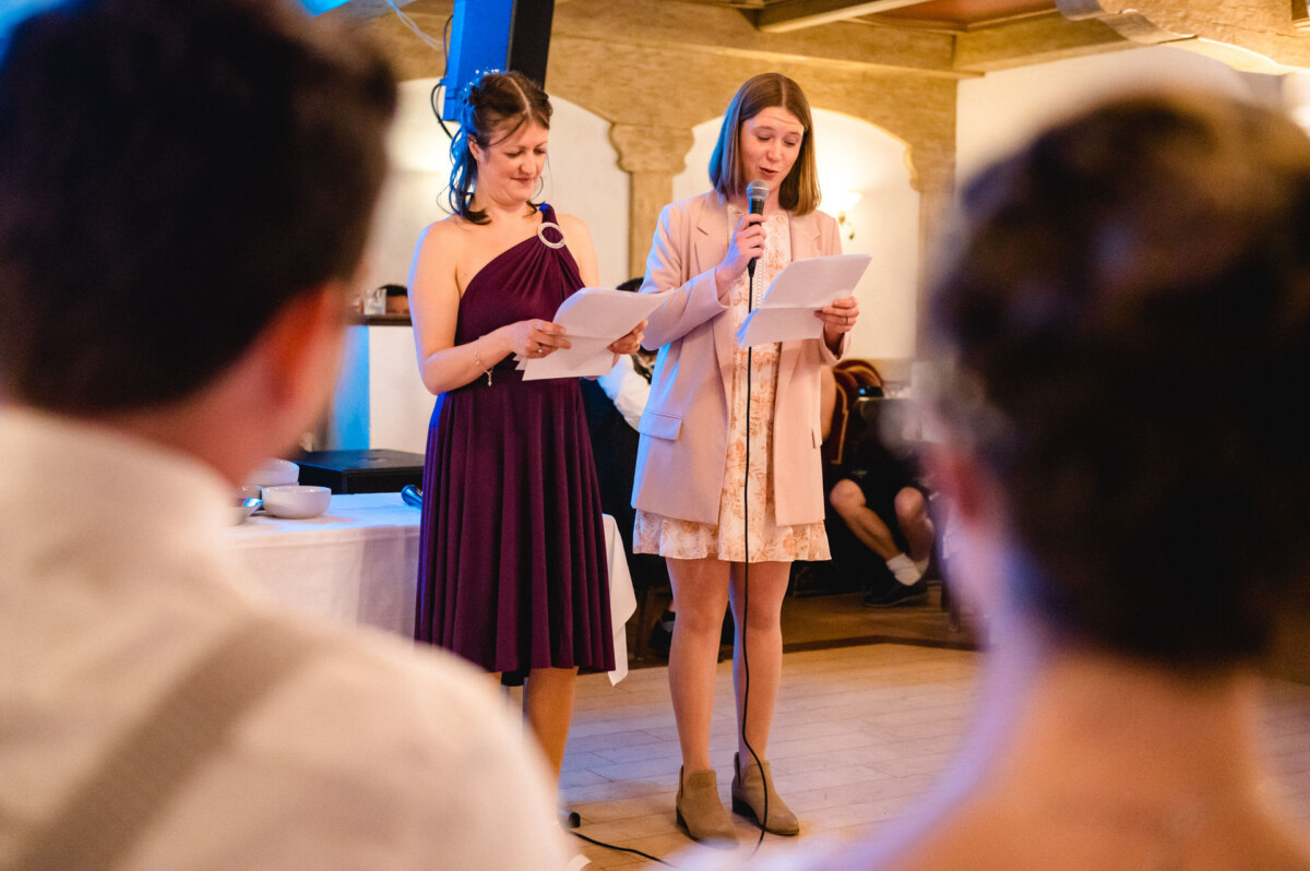 Zwei Frauen stehen drinnen und halten Papiere in der Hand. Eine trägt ein lila Kleid und hält ein Mikrofon; die andere trägt ein rosa Kleid und eine Jacke. Sie scheinen eine Rede zu halten. Im Vordergrund ist das Publikum zu sehen.