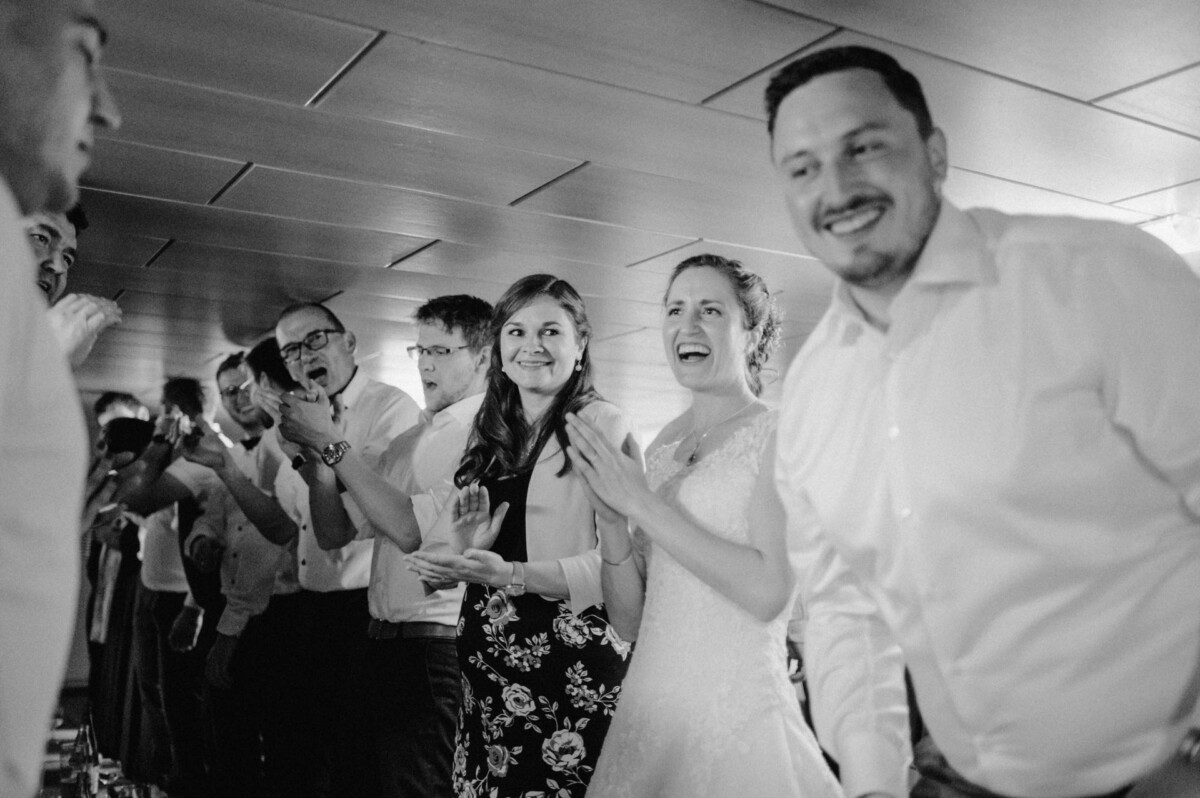 Eine Gruppe lächelnder und klatschender Menschen bei einer Feier; im Vordergrund steht ein Paar, die Frau trägt ein Brautkleid.