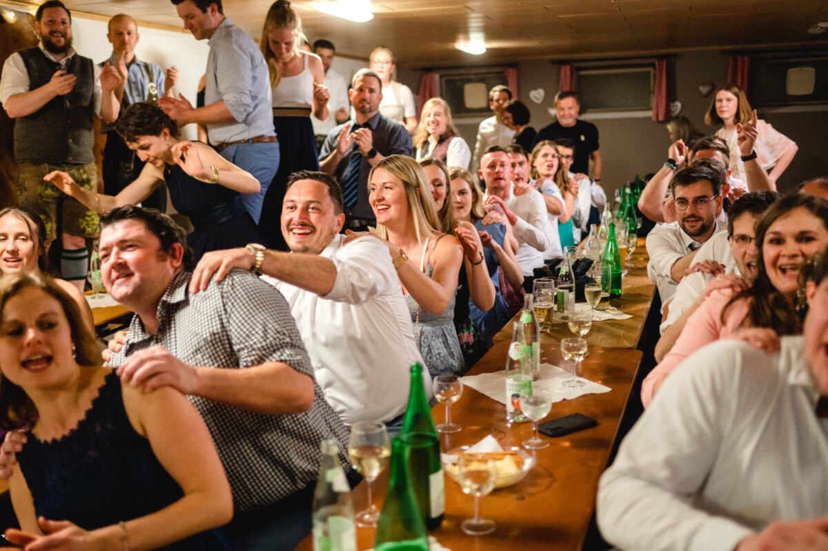 Eine Gruppe von Menschen sitzt dicht gedrängt an langen Tischen, genießt Getränke und Essen, viele lächeln und unterhalten sich. Auf den Tischen sind Flaschen und Gläser zu sehen.