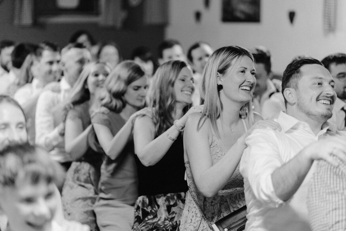 Bei einer gut besuchten Indoor-Veranstaltung stellt sich eine Gruppe von Menschen zum Conga-Tanz auf, lächelt und hat Spaß.