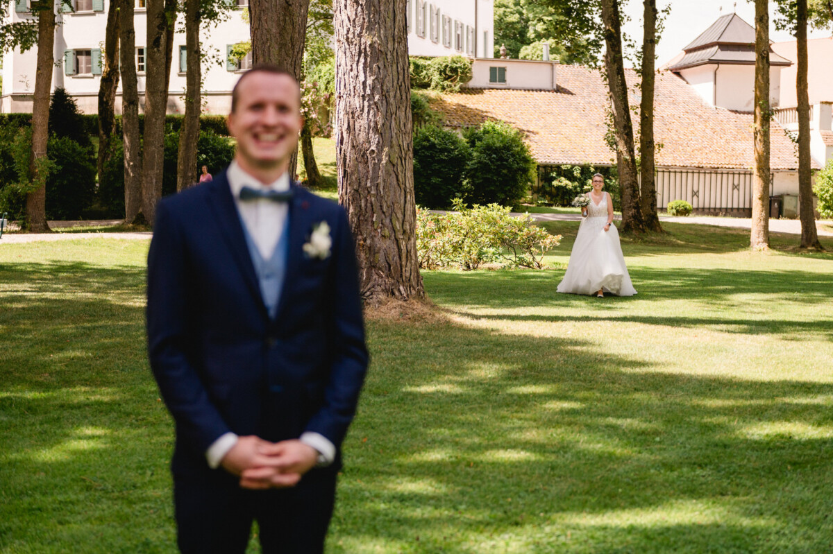 Im Vordergrund steht lächelnd ein Bräutigam im marineblauen Anzug, während in einer Parkanlage eine Braut im weißen Kleid auf ihn zugeht.