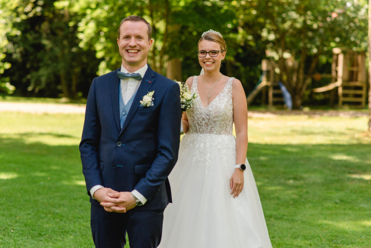 Braut und Bräutigam stehen draußen auf einer Rasenfläche. Der Bräutigam trägt einen dunklen Anzug und die Braut ein weißes Kleid und hält einen kleinen Blumenstrauß in der Hand. Sie lächeln und scheinen bereit für einen formellen Anlass zu sein.
