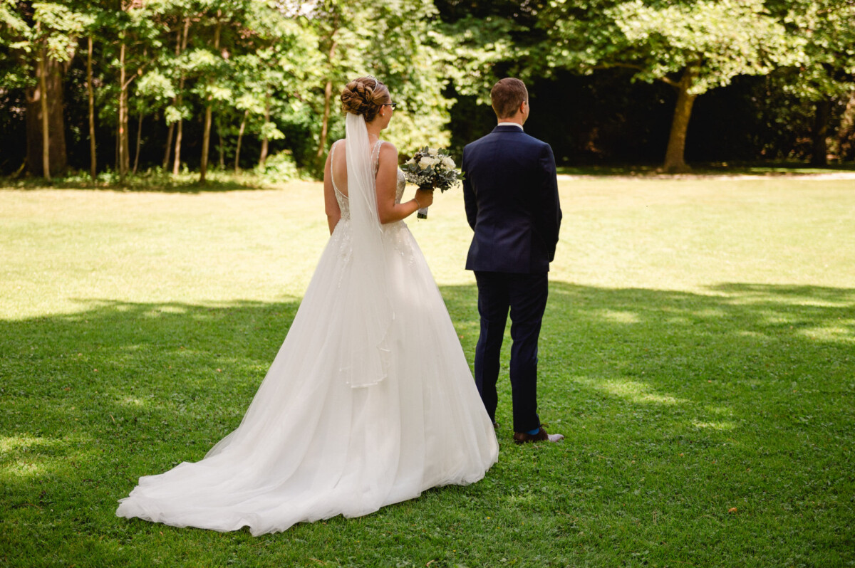 Eine Braut in einem weißen Kleid und Schleier steht mit Blumen in der Hand neben einem Bräutigam im marineblauen Anzug auf einer Wiese mit Bäumen im Hintergrund.