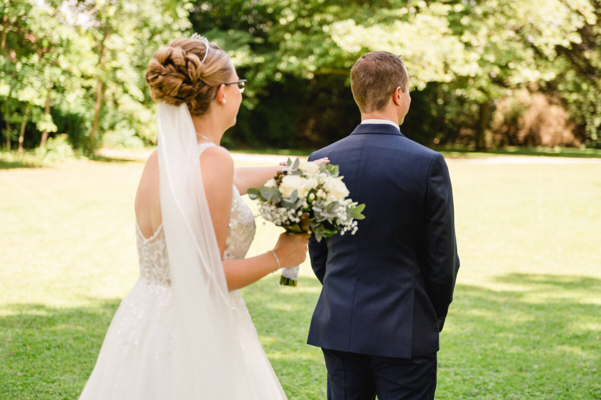 Eine Braut mit einem Blumenstrauß nähert sich einem Bräutigam, der mit dem Rücken zu einer Grasfläche steht.