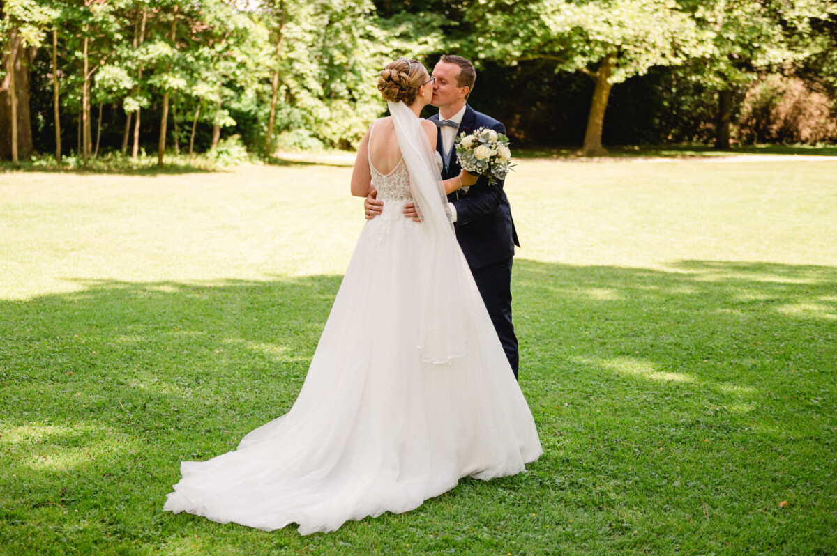 Ein Paar im Hochzeitskleid umarmt und küsst sich auf einer Wiese, umgeben von Bäumen.