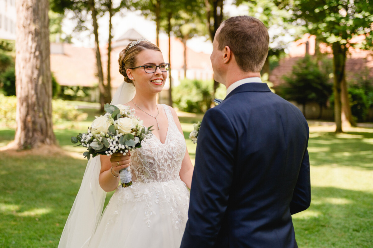 Braut und Bräutigam lächeln sich in einer sonnigen Umgebung im Freien mit Bäumen an. Die Braut hält einen Blumenstrauß und trägt eine Brille und eine Tiara.