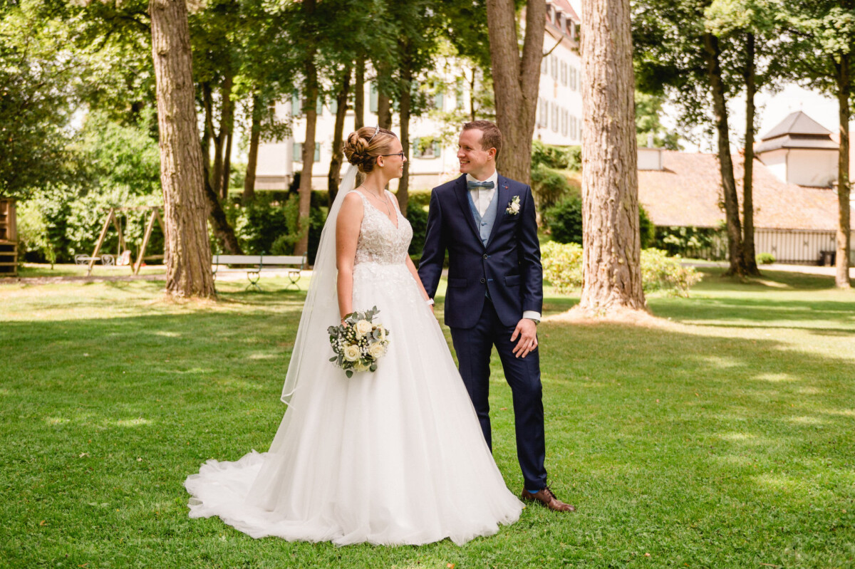 Braut und Bräutigam stehen auf einer von Bäumen umgebenen Wiese und blicken einander an. Die Braut hält einen Blumenstrauß und beide tragen Hochzeitskleidung.