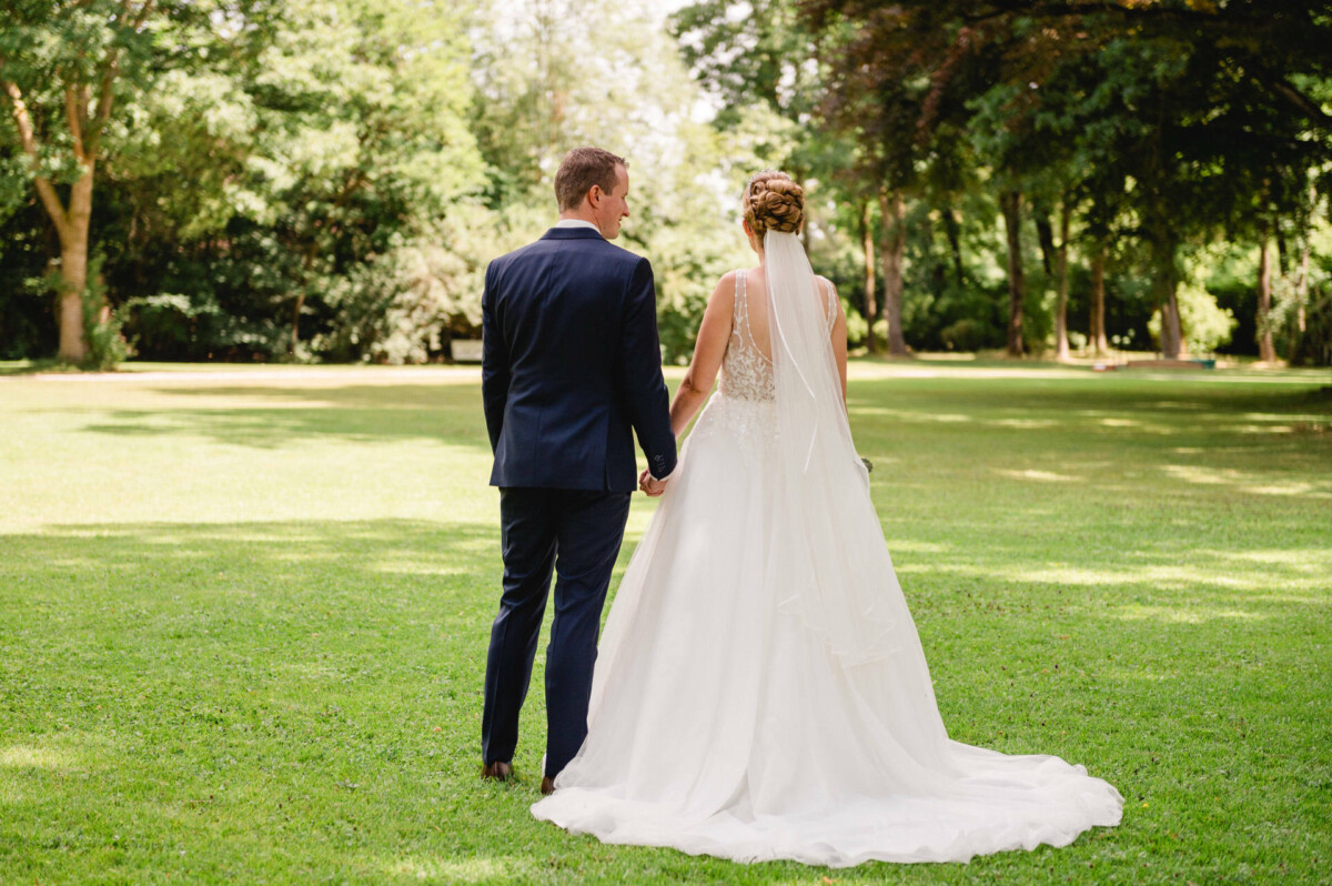 Braut und Bräutigam stehen auf dem Gras und halten sich an den Händen, im Hintergrund sind Bäume zu sehen.