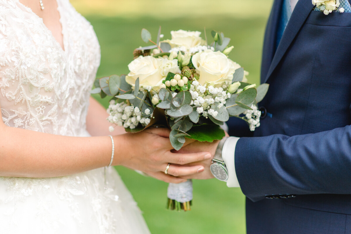 Eine Braut und ein Bräutigam stehen im Freien und halten einen Strauß weißer Rosen und Grünpflanzen in der Hand.