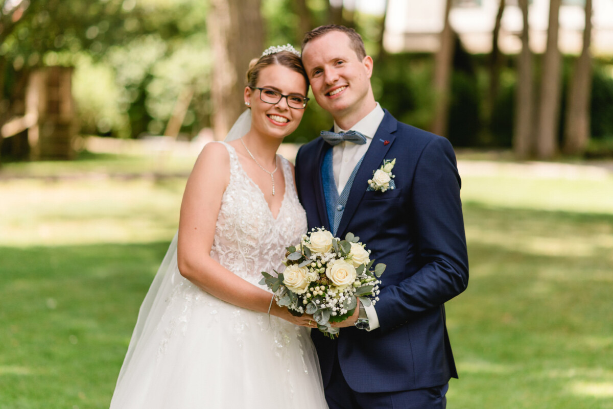 Braut und Bräutigam stehen zusammen im Freien, lächeln und halten einen Strauß weißer Rosen in den Händen. Die Braut trägt ein weißes Kleid und einen Schleier, der Bräutigam einen marineblauen Anzug mit Fliege.