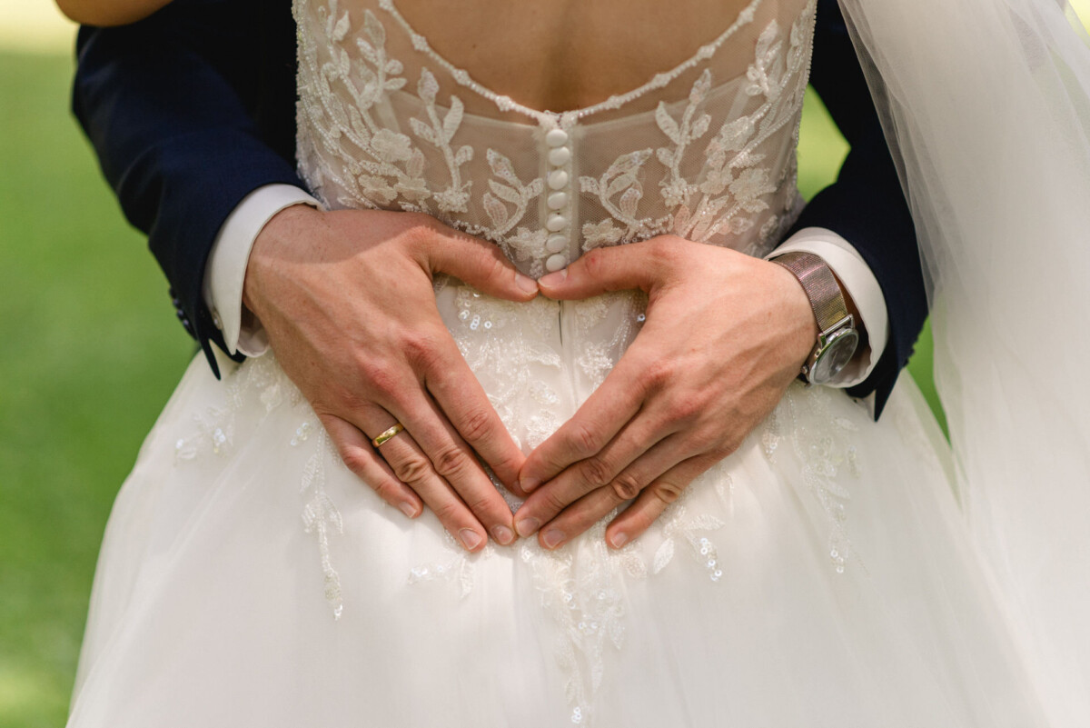 Nahaufnahme der Hände eines Bräutigams, die über dem Rücken des Spitzenhochzeitskleides einer Braut ein Herz formen.