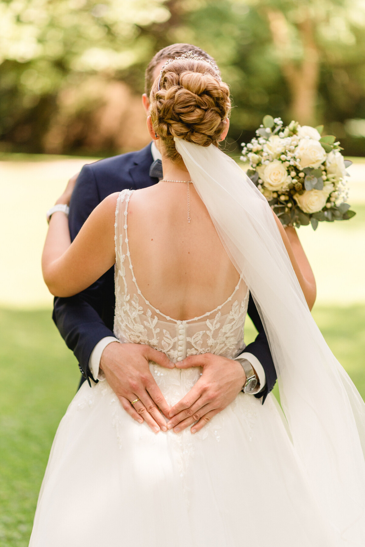 Braut und Bräutigam umarmen sich in einem Garten. Die Braut hält einen Strauß weißer Rosen in der Hand. Der Bräutigam formt mit seinen Händen auf dem Rücken der Braut ein Herz.