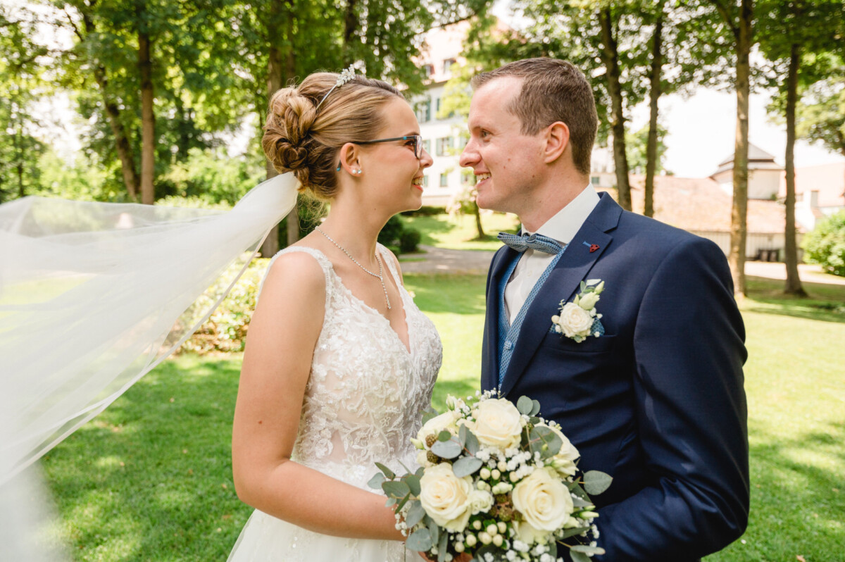 Eine Braut und ein Bräutigam lächeln sich in einem Park an. Die Braut hält einen Strauß weißer Blumen in der Hand und beide tragen Hochzeitskleidung.
