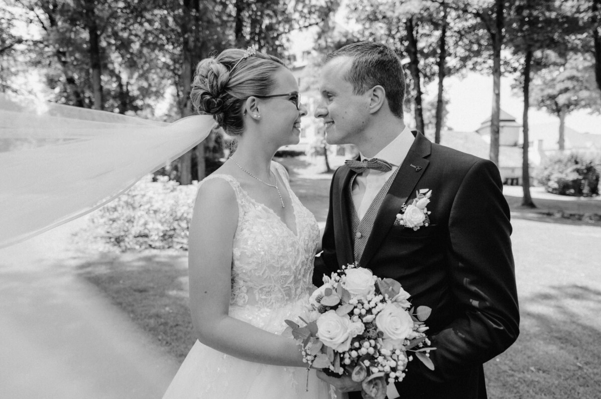 Braut und Bräutigam lächeln sich im Freien, umgeben von Bäumen, an. Die Braut hält einen Blumenstrauß und ihr Schleier weht im Wind. Schwarzweißfoto.