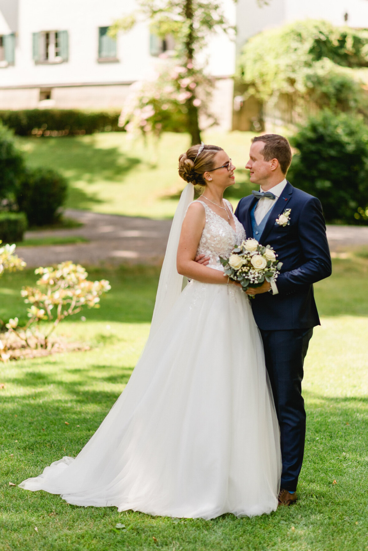 Braut und Bräutigam stehen auf dem Gras in einem Garten, stehen einander gegenüber und lächeln. Die Braut hält einen Blumenstrauß.