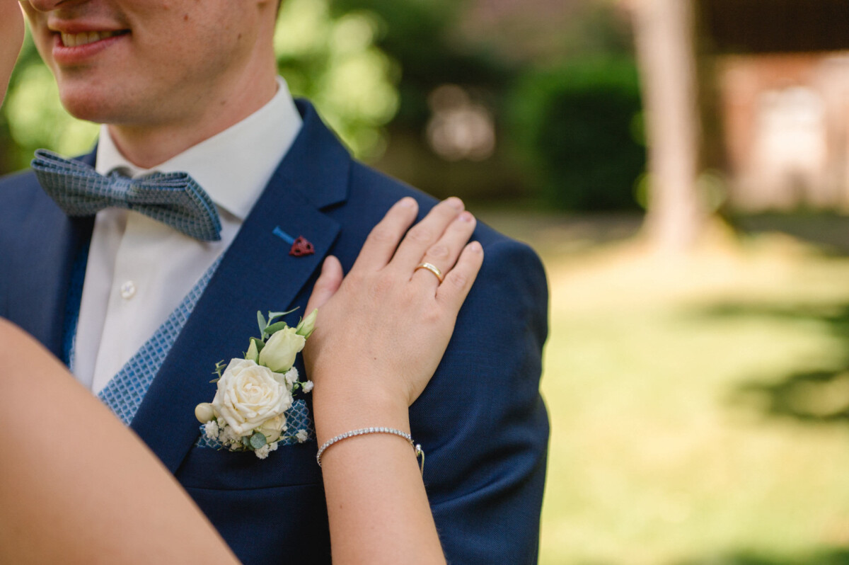 Eine Person in einem blauen Anzug mit Fliege und Ansteckblume aus einer weißen Rose, mit einer Hand auf der Schulter im Freien.