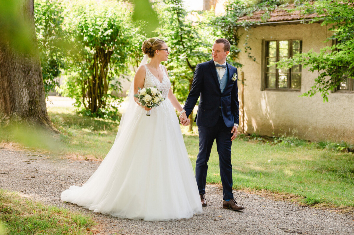 Braut und Bräutigam gehen Hand in Hand einen Kiesweg entlang, umgeben von Grün. Die Braut hält einen Blumenstrauß und beide tragen formelle Hochzeitskleidung.