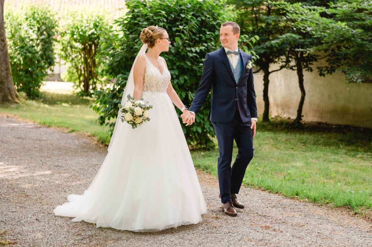 Braut und Bräutigam gehen Hand in Hand auf einem von Grün umgebenen Kiesweg. Die Braut hält einen Blumenstrauß.