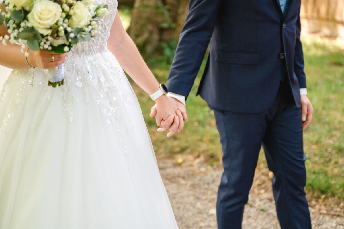 Eine Braut in einem weißen Kleid und ein Bräutigam in einem dunklen Anzug halten sich in einer Gartenkulisse an den Händen. Die Braut hält einen Strauß weißer Rosen.