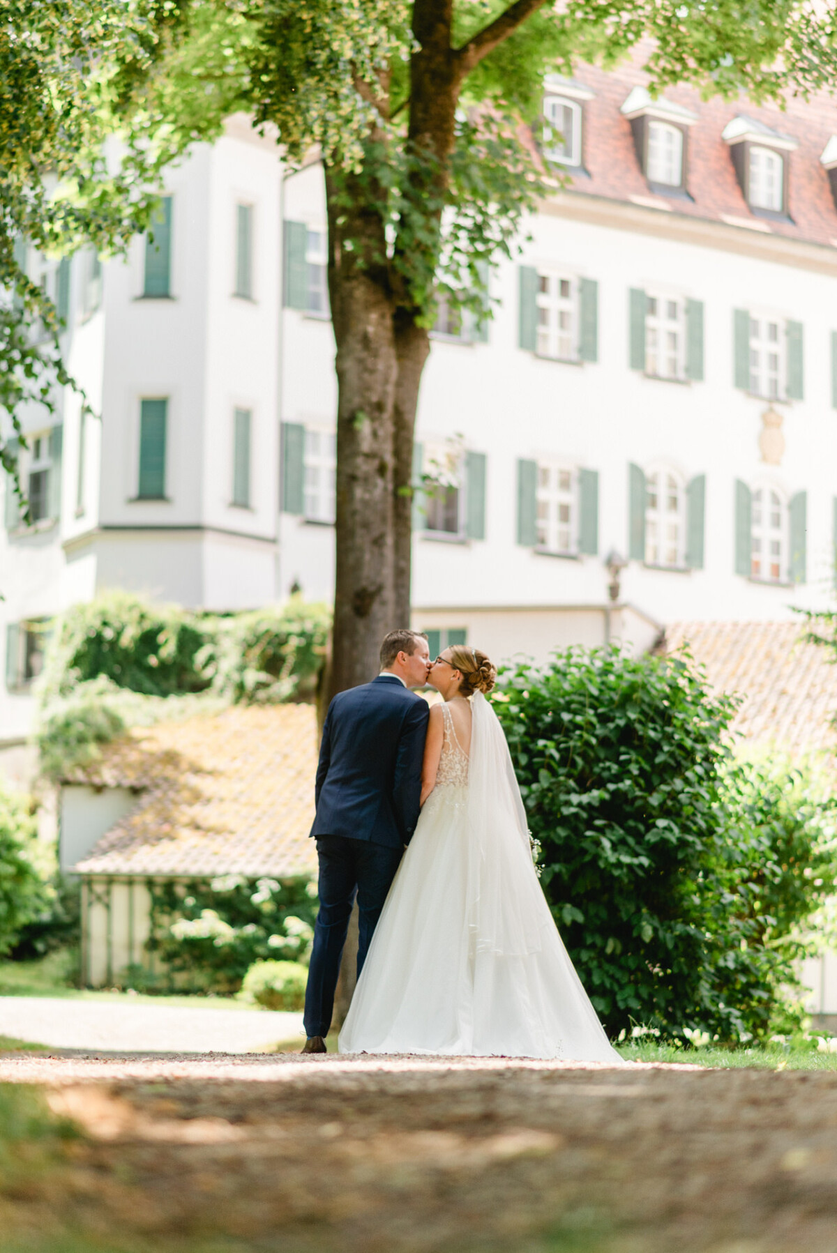 Ein Paar im Hochzeitskleid steht unter einem Baum und küsst sich. Im Hintergrund ist ein großes Gebäude zu sehen.