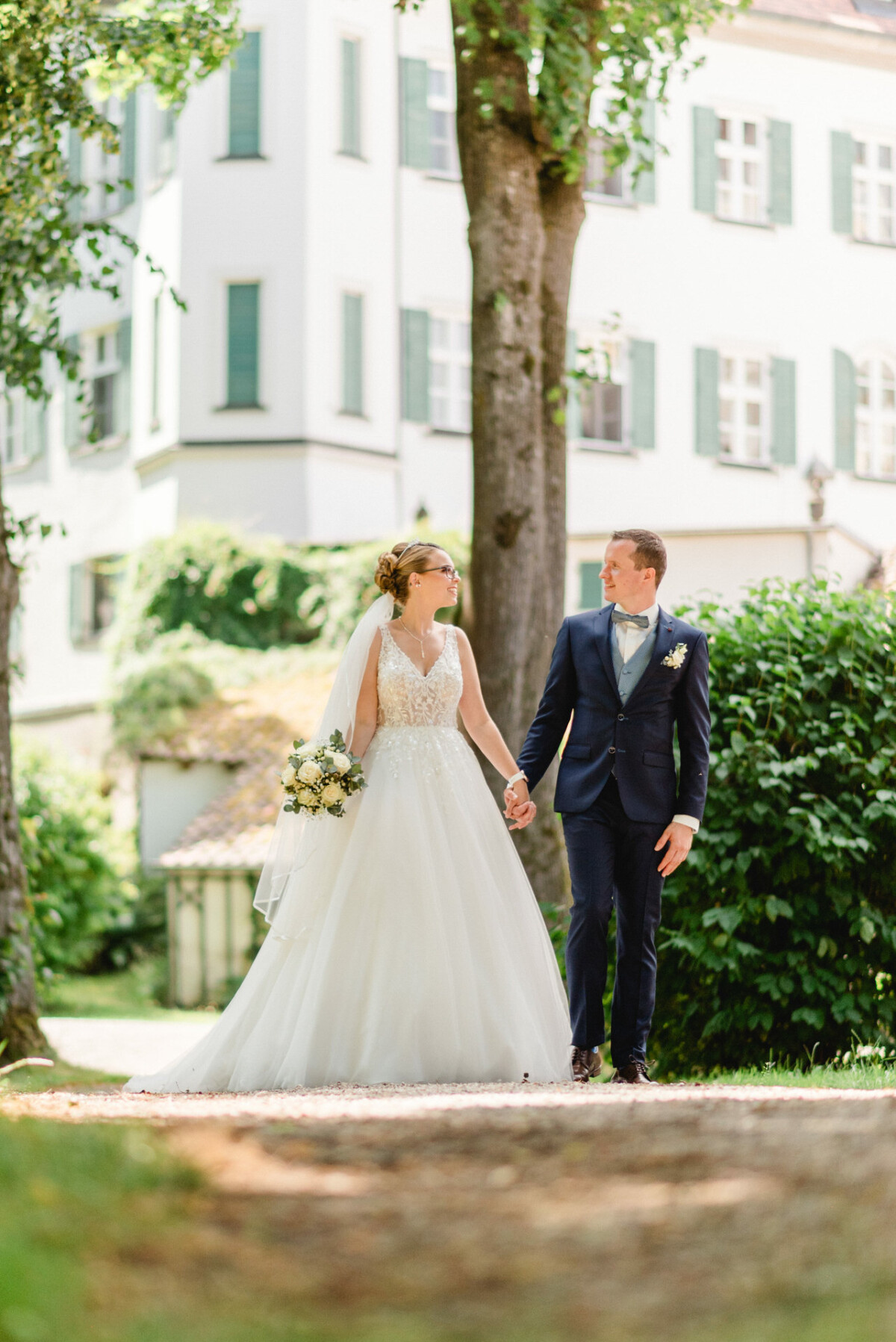 Braut und Bräutigam gehen Händchen haltend im Freien spazieren. Sie trägt ein weißes Kleid und hält einen Blumenstrauß, während er einen dunklen Anzug trägt. Im Hintergrund sind Bäume und ein Gebäude zu sehen.