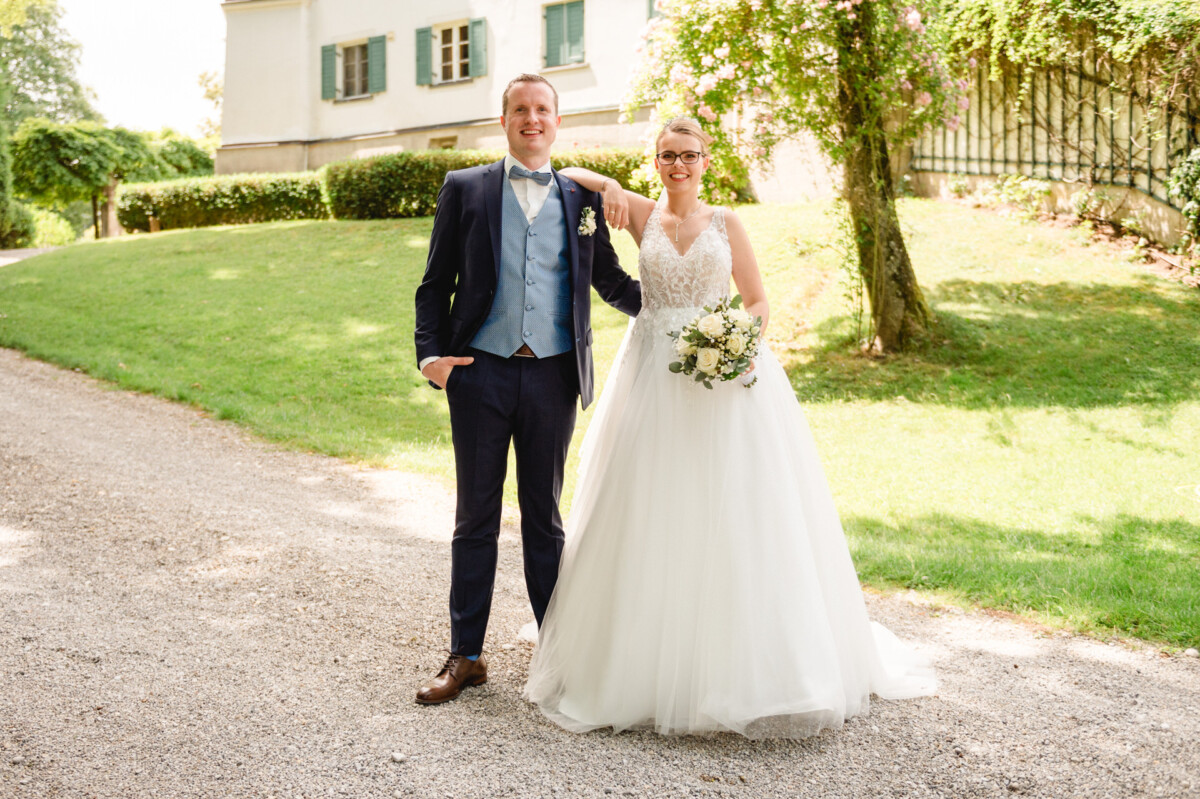 Eine Braut und ein Bräutigam posieren zusammen an einem sonnigen Tag. Sie stehen auf einem Kiesweg mit viel Grün und einem Gebäude im Hintergrund.