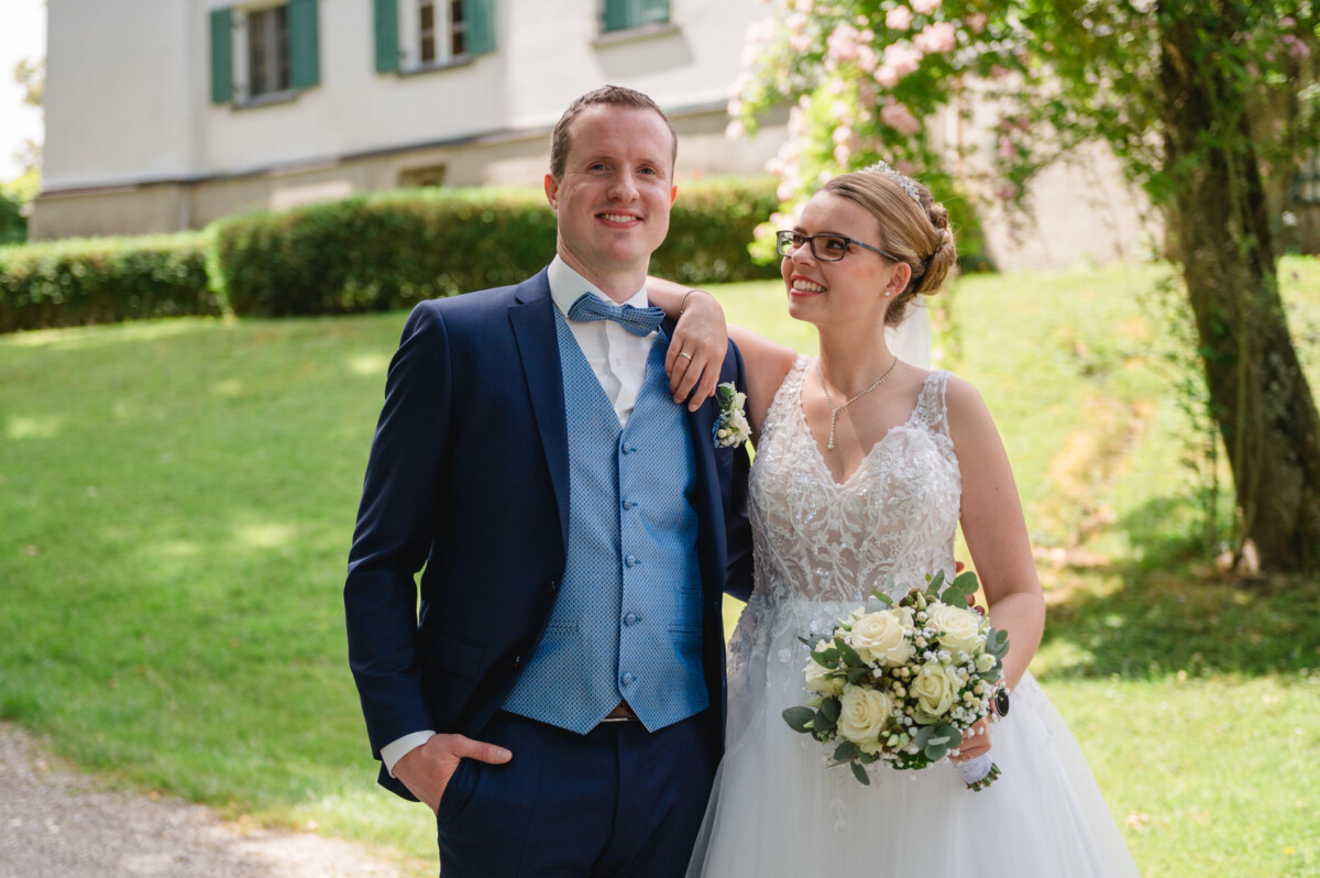 Braut und Bräutigam lächeln im Freien, die Braut hält einen Blumenstrauß. Sie tragen formelle Hochzeitskleidung und stehen auf einem Gartenweg mit einem Gebäude und viel Grün im Hintergrund.