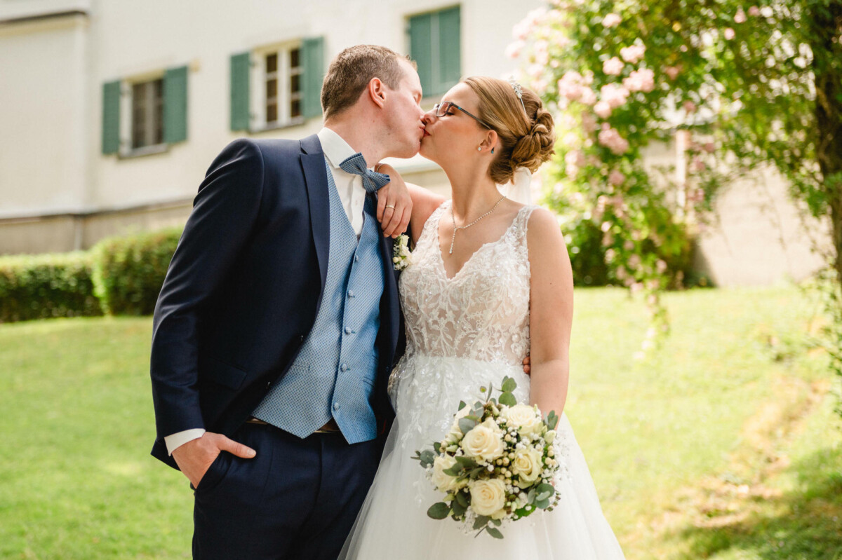 Braut und Bräutigam küssen sich draußen vor einem Haus. Die Braut hält einen Strauß weißer Blumen in der Hand und beide tragen Abendgarderobe.