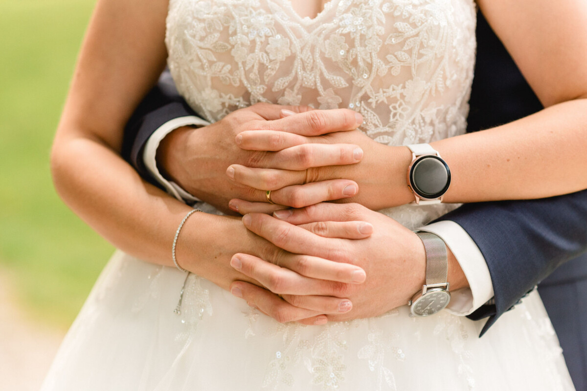 Ein Paar umarmt sich, von den Schultern abwärts sichtbar. Die Braut trägt ein Spitzenkleid und der Bräutigam einen dunklen Anzug. Beide tragen Uhren.