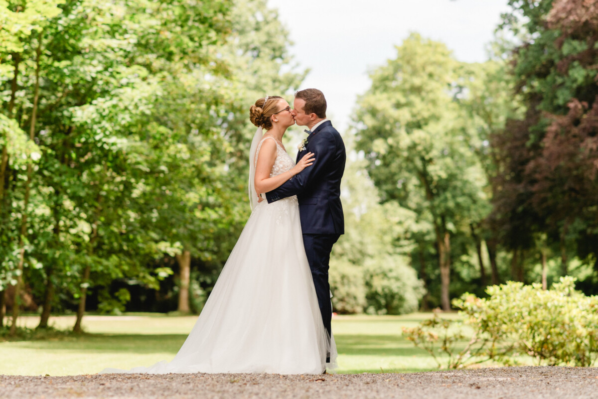 Braut und Bräutigam küssen sich im Freien in einer Gartenanlage, umgeben von Grün.