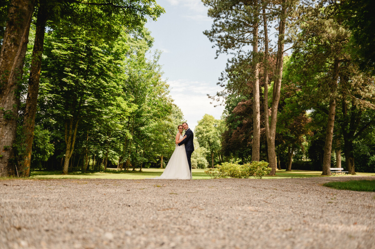 Braut und Bräutigam umarmen sich auf einem Kiesweg in einer üppigen Parklandschaft mit Bäumen und einer Picknickbank im Hintergrund.