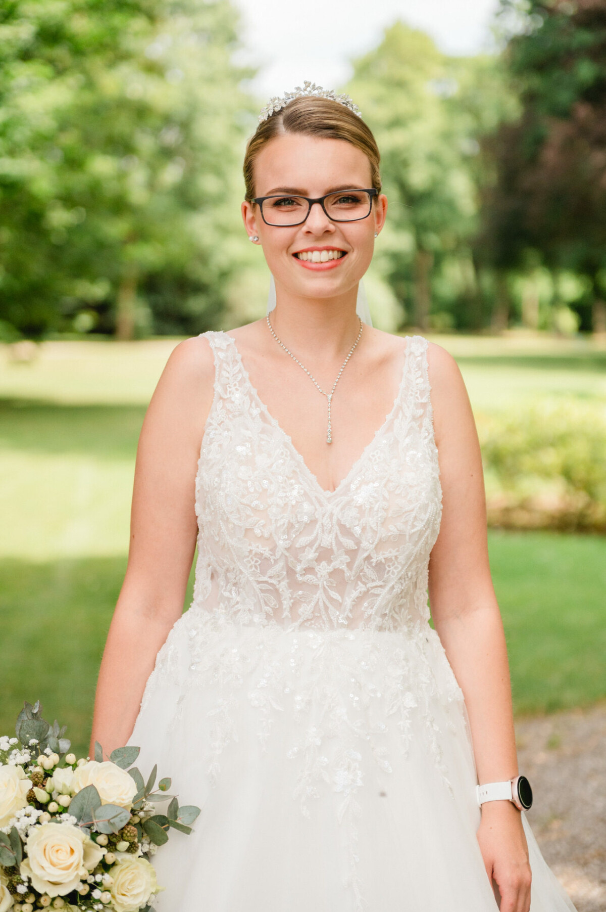 Eine Frau in einem weißen Spitzenbrautkleid und mit Brille steht draußen und hält einen Strauß weißer Rosen und Grünpflanzen in der Hand. Sie lächelt, im Hintergrund sind Bäume und Gras zu sehen.