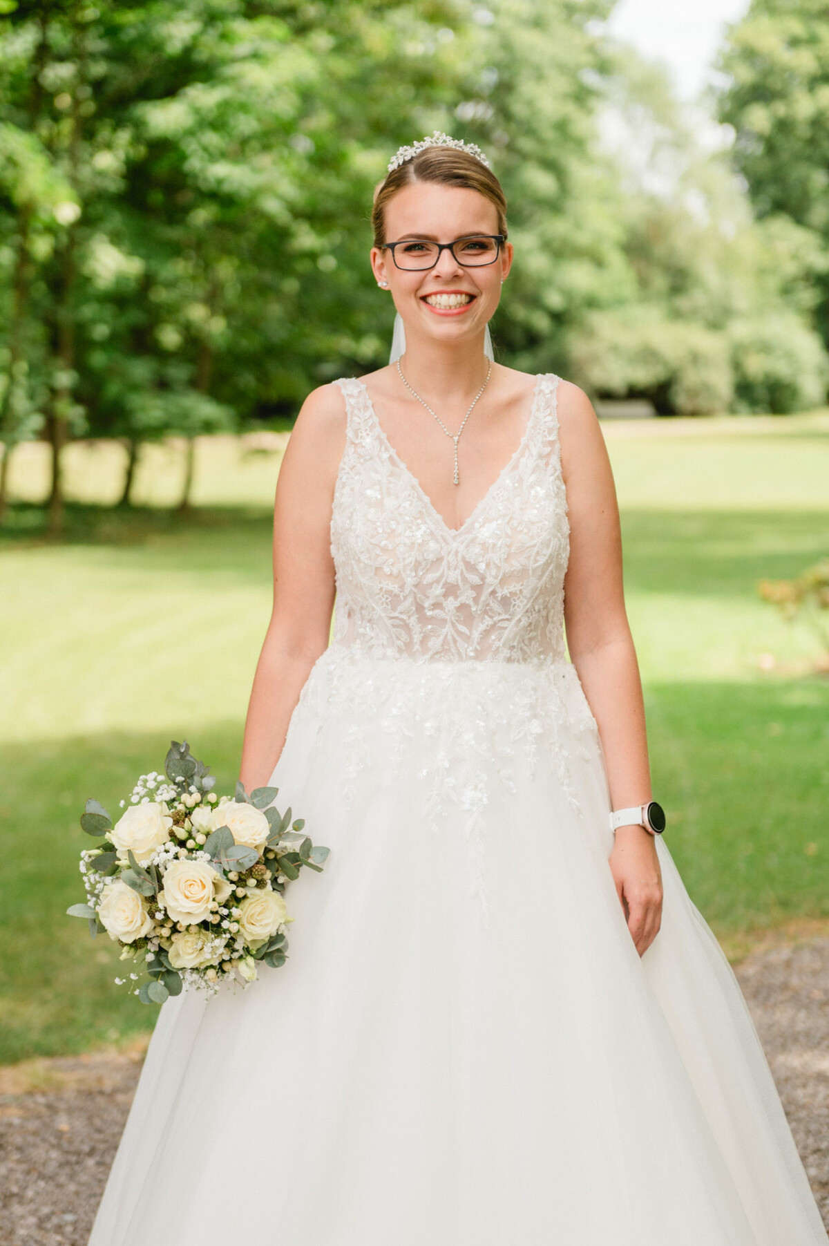 Eine Braut in einem weißen Spitzenkleid hält einen Strauß weißer Rosen und Grünpflanzen und steht draußen auf einem Gartenweg.