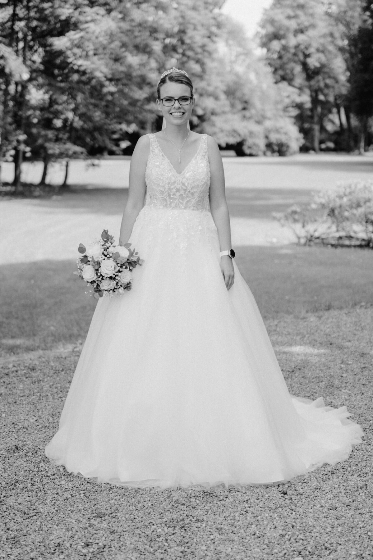 Eine Braut in einem weißen Brautkleid hält einen Blumenstrauß und steht auf einem Kiesweg in einer Gartenanlage.
